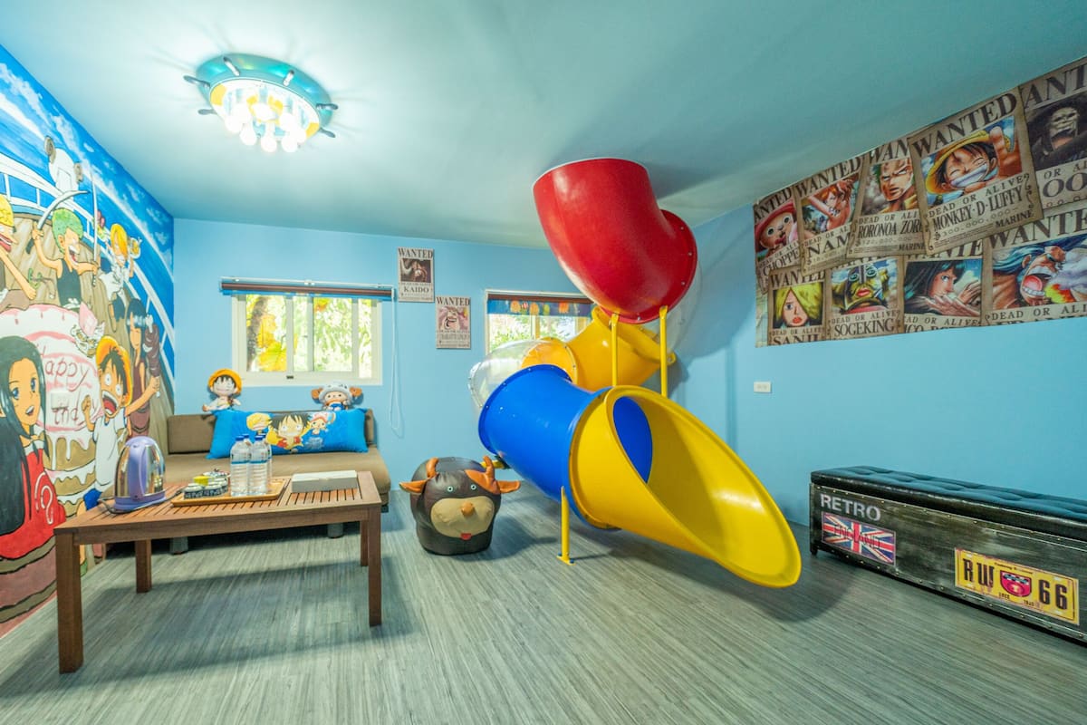 A room with a large mural, a play area with a slide, a sofa, a table with toys, and a window with a view of trees.