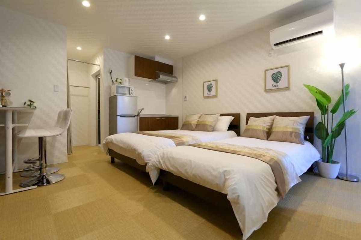 A hotel room with a bed, a desk, a chair, a potted plant, and a kitchenette.