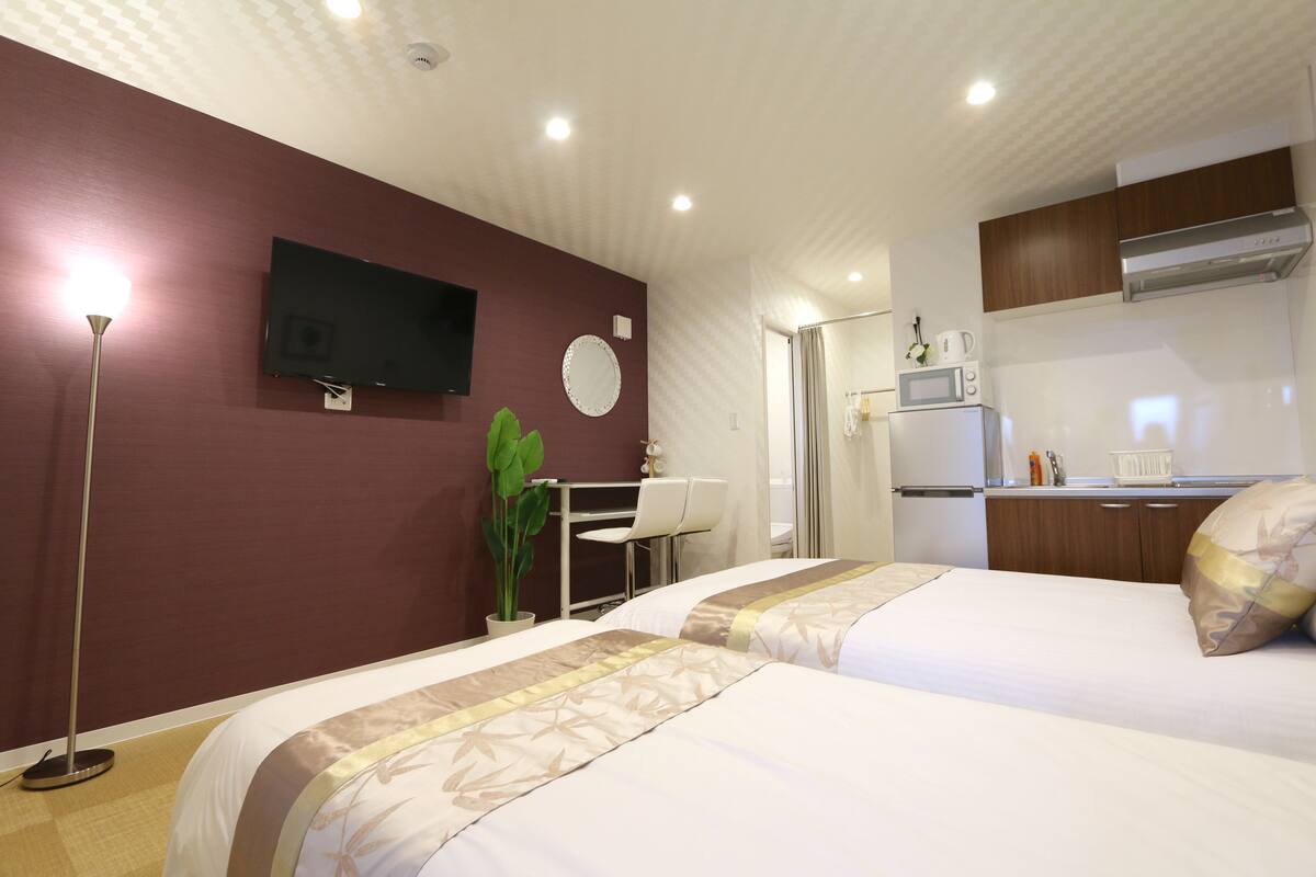 A hotel room with two beds, a TV, a desk with a chair, a small kitchenette, and a potted plant.