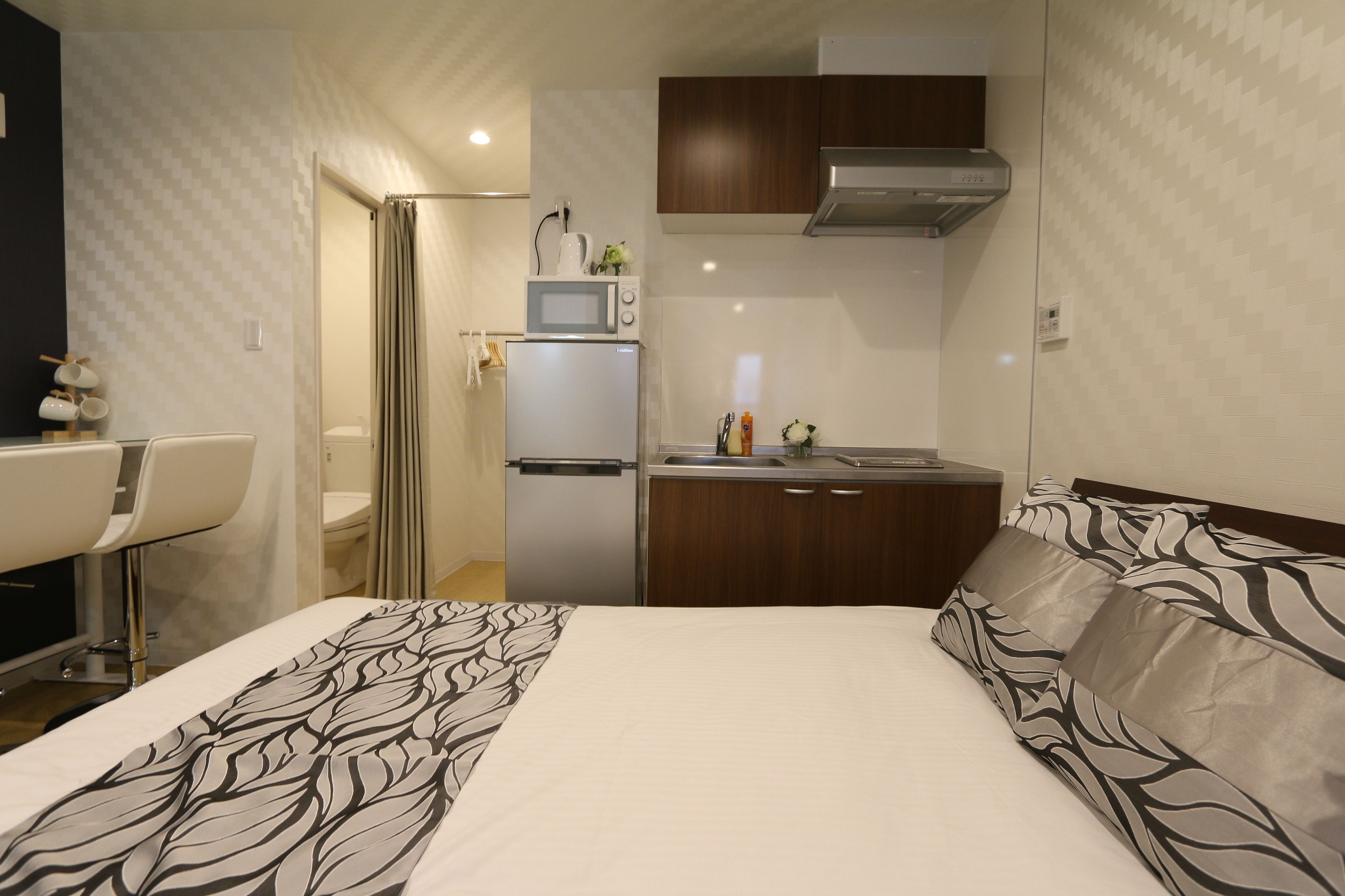 A compact hotel room with a bed, a small kitchenette, a bathroom, and a sitting area.