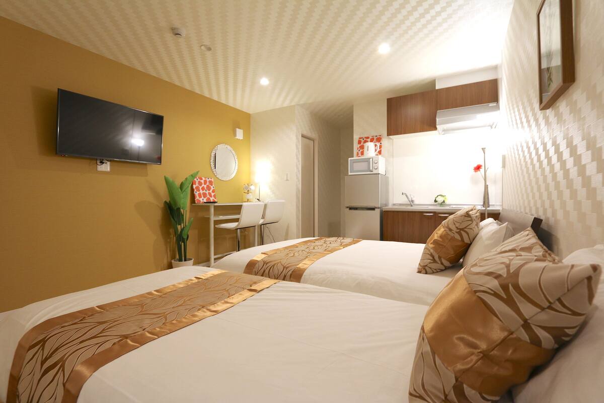 A hotel room with two beds, a TV, a desk with a chair, a microwave, and a small kitchenette.