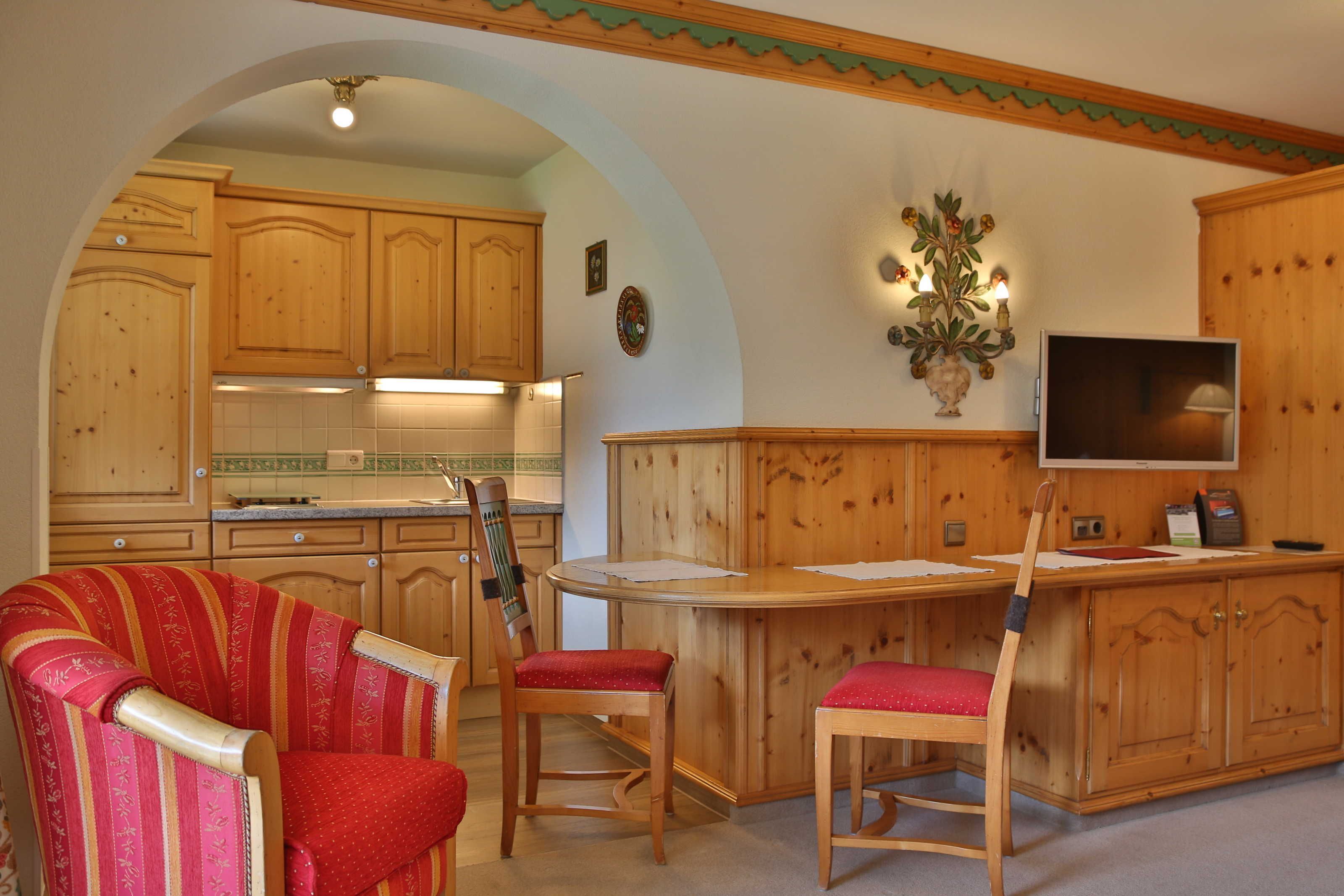 A kitchen with wooden cabinets, a dining area with a red upholstered chair, and a television on a wooden cabinet.