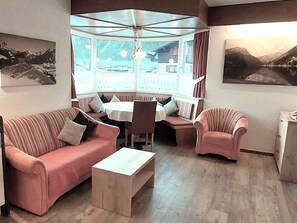 Un ambiente accogliente con divano, poltrona e un tavolino. Si vede una finestra con tende, una ventola a soffitto e un quadro appeso al muro.