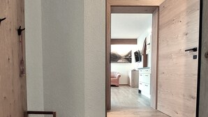 A hallway leading to a room with a wooden floor, a bench, and a view of a kitchen with a chair and cabinets.