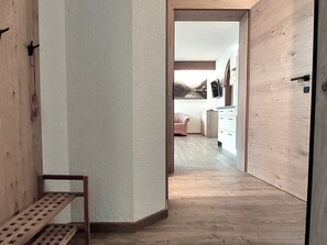 Un couloir mène à une pièce dotée d’un plancher en bois, d’un banc et d’une vue sur une cuisine avec une chaise et des placards.