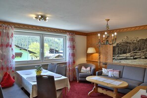 Ein Zimmer mit Esstisch, einer Couch, einem Sessel, einer Chandelüster und einem fenster mit Vorhängen.
