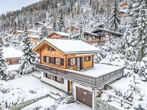 A wooden chalet with a balcony, surrounded by snow-covered trees and a mountainous backdrop.