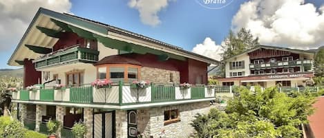 Un edificio de dos plantas con balcones, un coche aparcado delante y montañas al fondo.