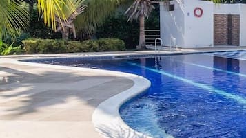 Piscina com borda curva, cercada por palmeiras e um prédio branco com um círculo vermelho.