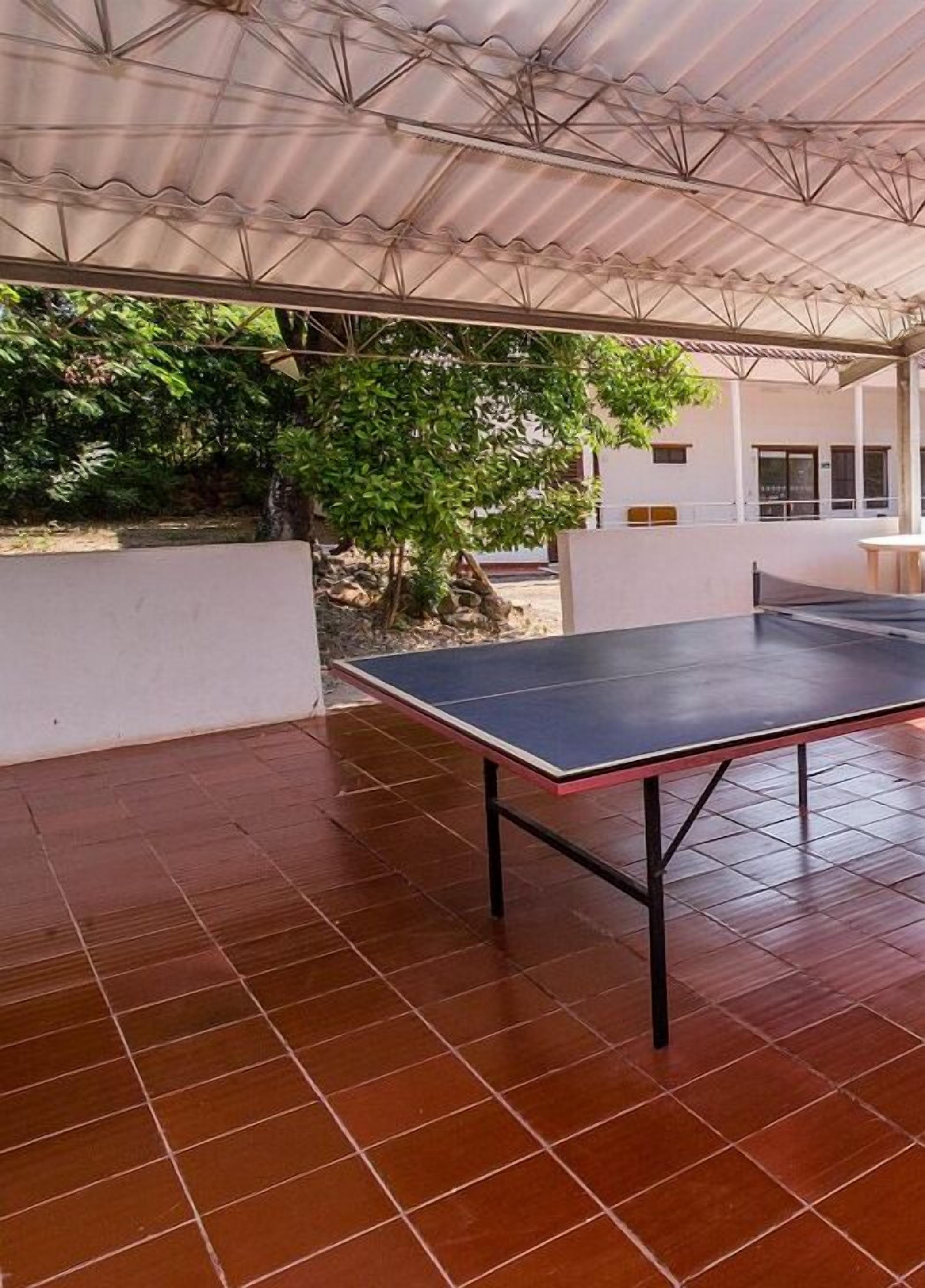 Un espacio al aire libre con mesa de ping pong, piso de baldosas y una estructura cubierta con techo de láminas.