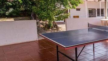 An outdoor area with a ping pong table, a tiled floor, and a covered structure with a corrugated roof.