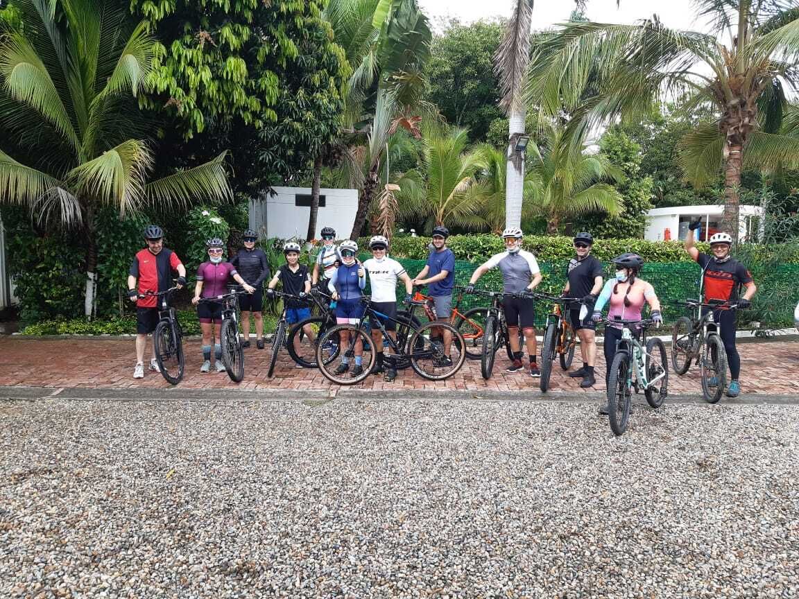 A group of cyclists standing in a line with bicycles, in a setting with palm trees and greenery.