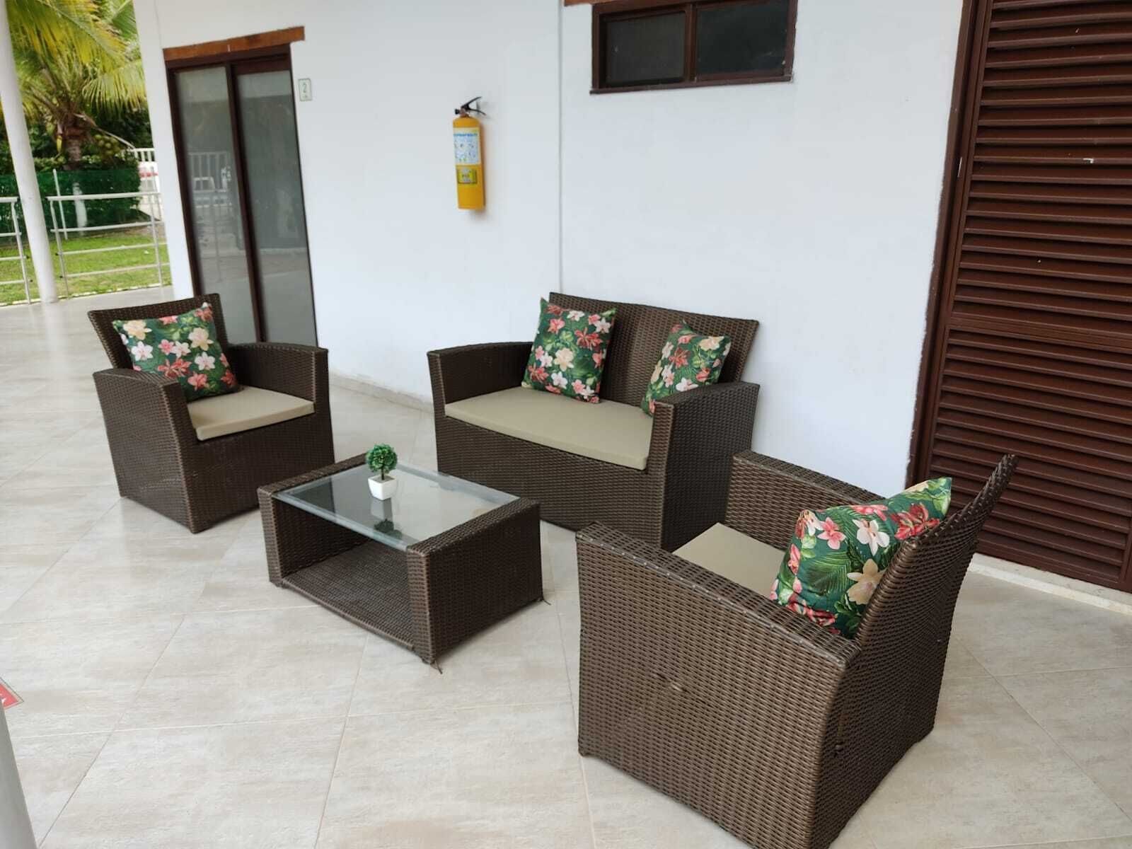 Uma área de pátio com móveis de vime, incluindo dois sofás e uma mesa de centro, e um extintor de incêndio fixado na parede.