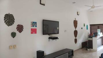 Una sala de estar con un televisor de pantalla plana montado en la pared, un estante pequeño debajo y un escritorio con una computadora y otros objetos.