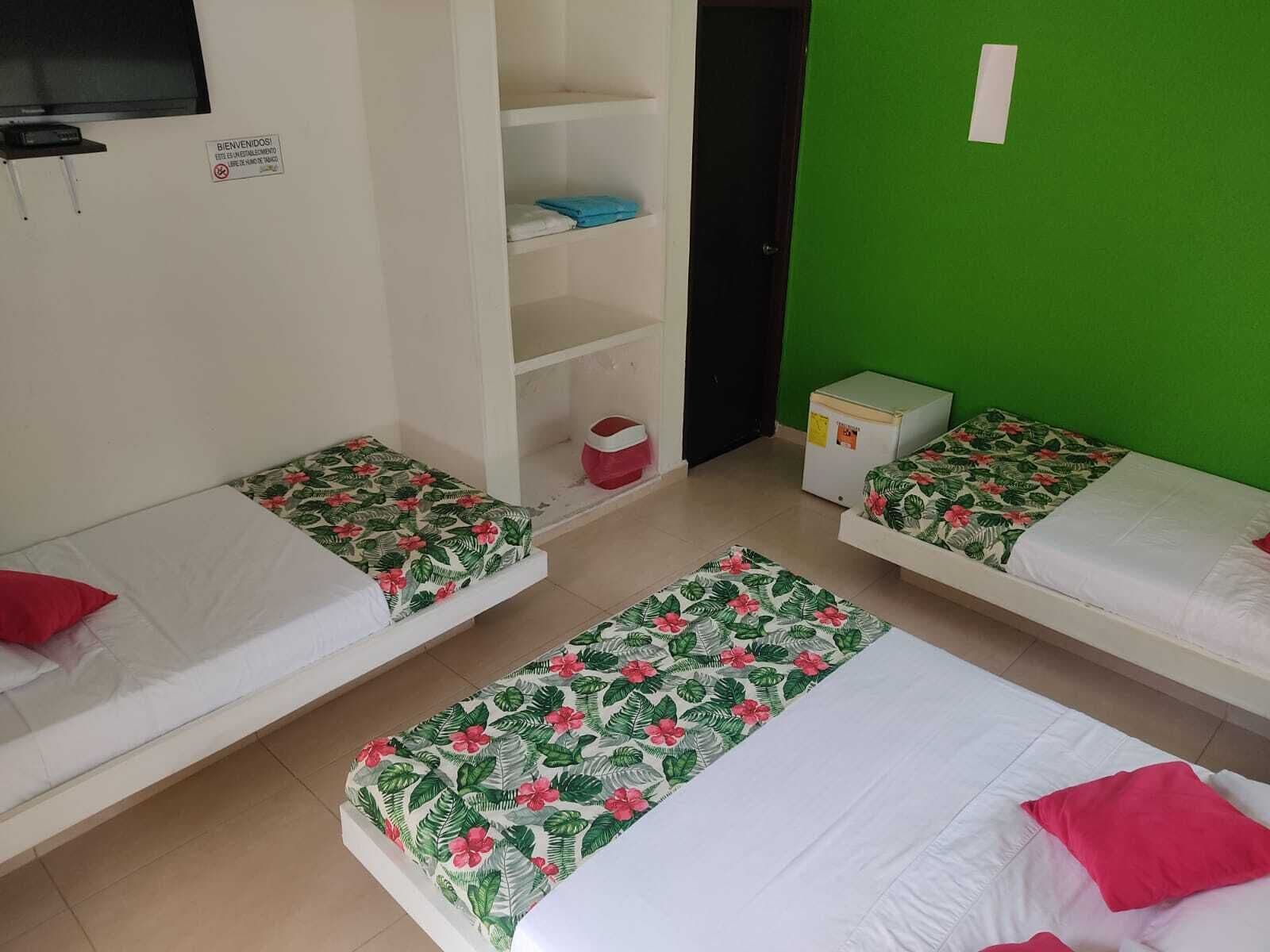 A room with three single beds, each with a floral bedspread and a red pillow. There is a small white shelf unit with folded clothes and a red basket. A white refrigerator is against the green wall.