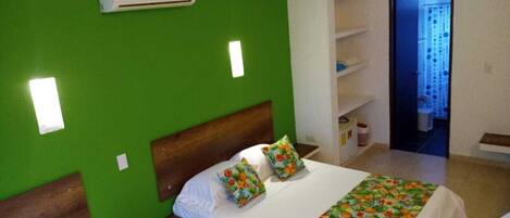 Un dormitorio con pared verde, cabecera de madera, cama blanca con almohadas florales, armario blanco y un baño pequeño visible a través de una puerta abierta.