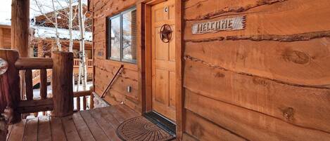 A wooden cabin with a wooden door marked "MELTINE," a window, and a porch with a bench.