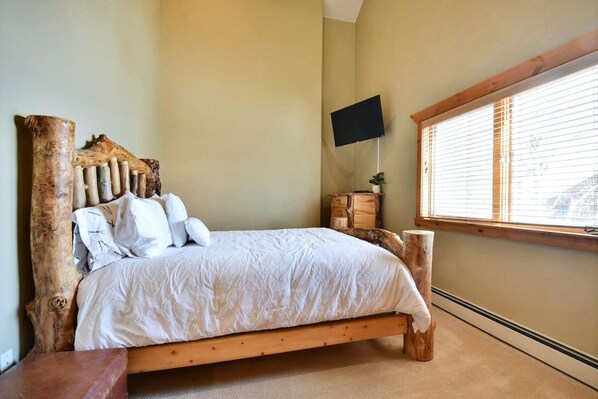 A bedroom with a wooden bed frame, a mounted TV, and a window with blinds.