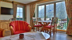 Une pièce avec une télévision, une table à manger avec des chaises, une fenêtre avec des rideaux et une vue sur les montagnes.