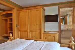 Habitación con muebles de madera, una cama con ropa de cama floral, un televisor y un baño visible a través de una puerta abierta.