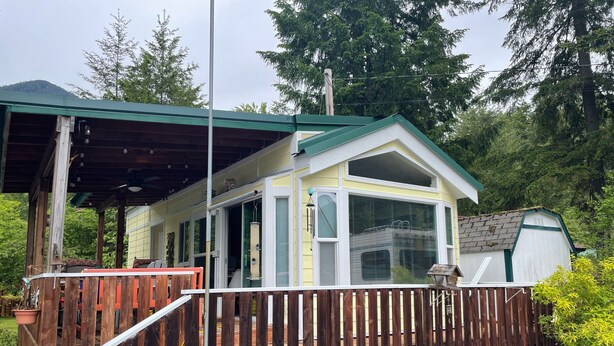 A wooden deck with a covered area, a fenced yard, and a house with a green roof and large windows. - Mountain Frog Cabin - A.C./Sleeps 2/Hot Tub - 30 min. from Rainier/Skiing (Packwood)
