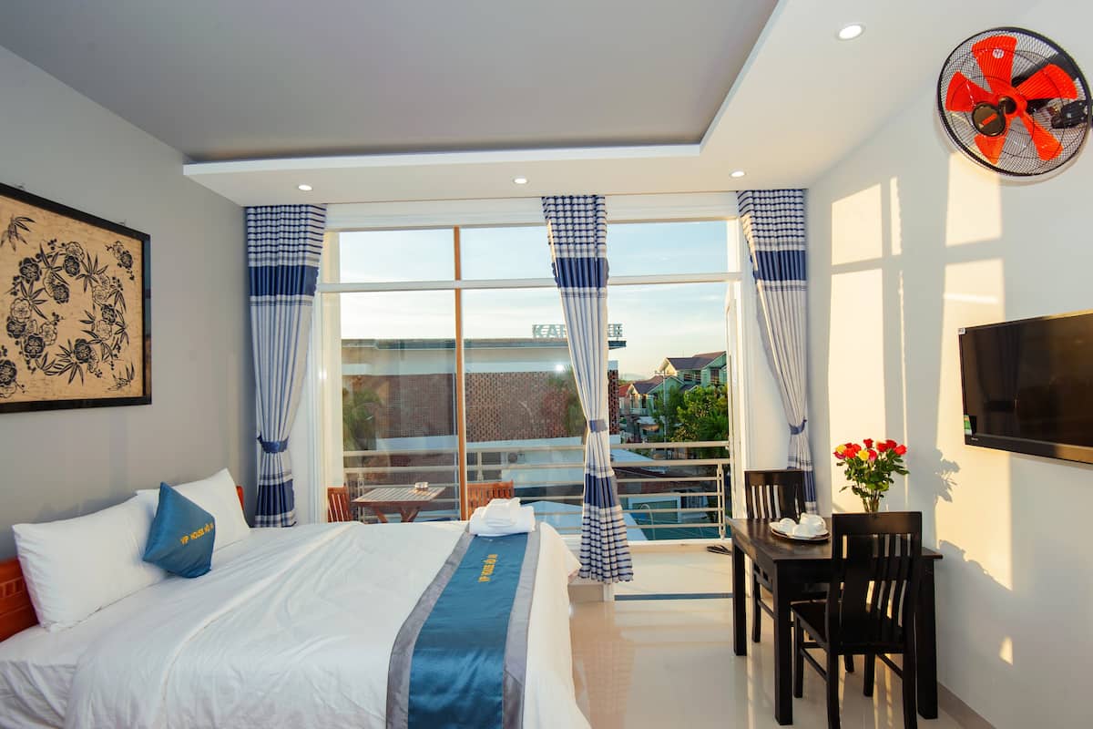 A hotel room with a bed, a dining table with chairs, a TV, and a balcony with curtains.