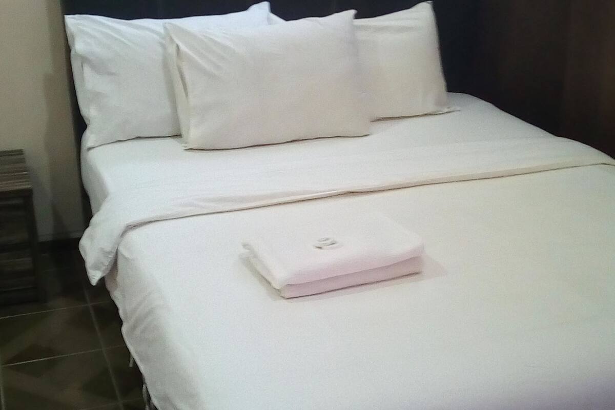 A neatly made bed with white bedding and pillows, a small bedside table, and a wall-mounted picture.