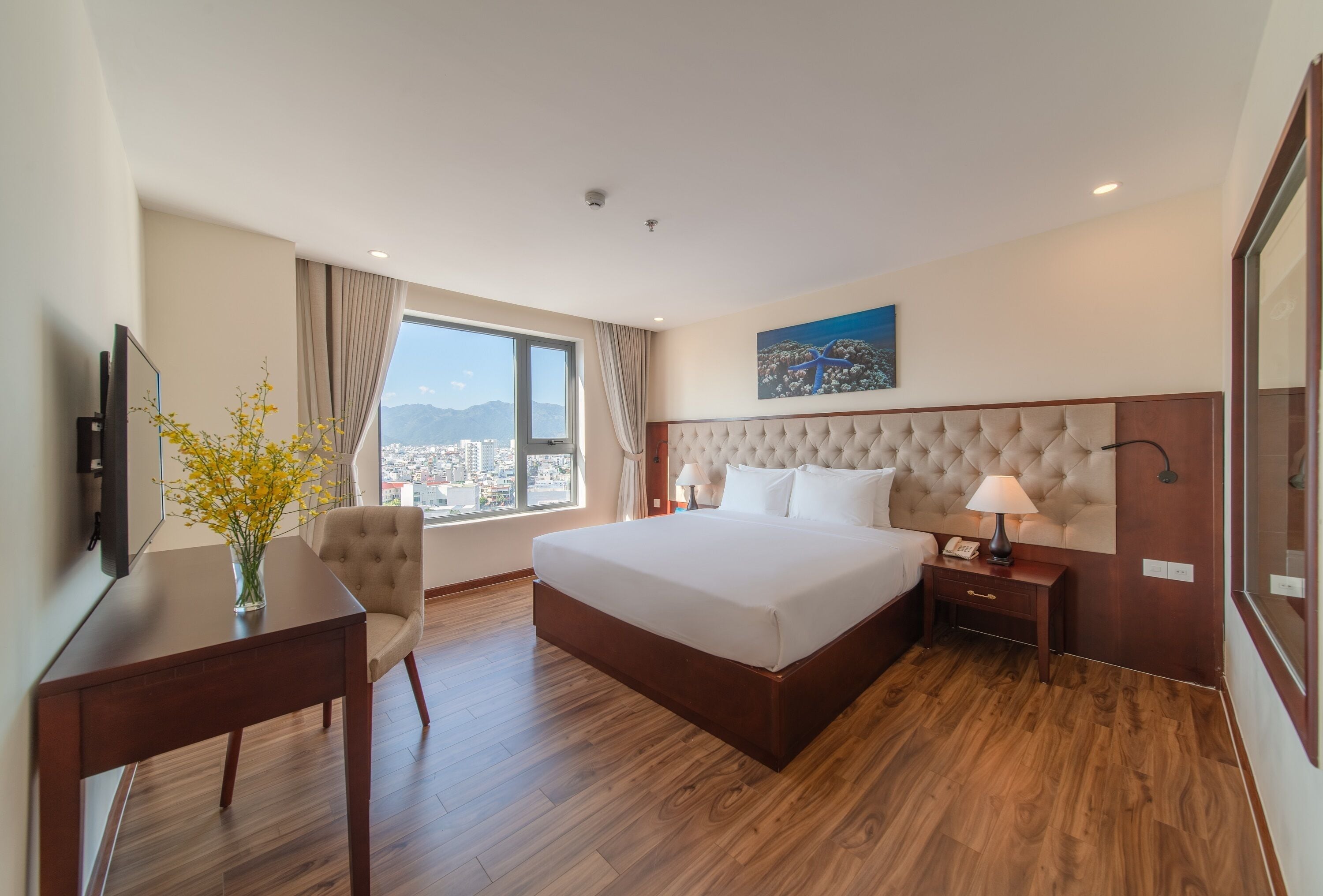 A hotel room with a large bed, a desk, a chair, a TV, a window with a view of the city, and a painting on the wall.