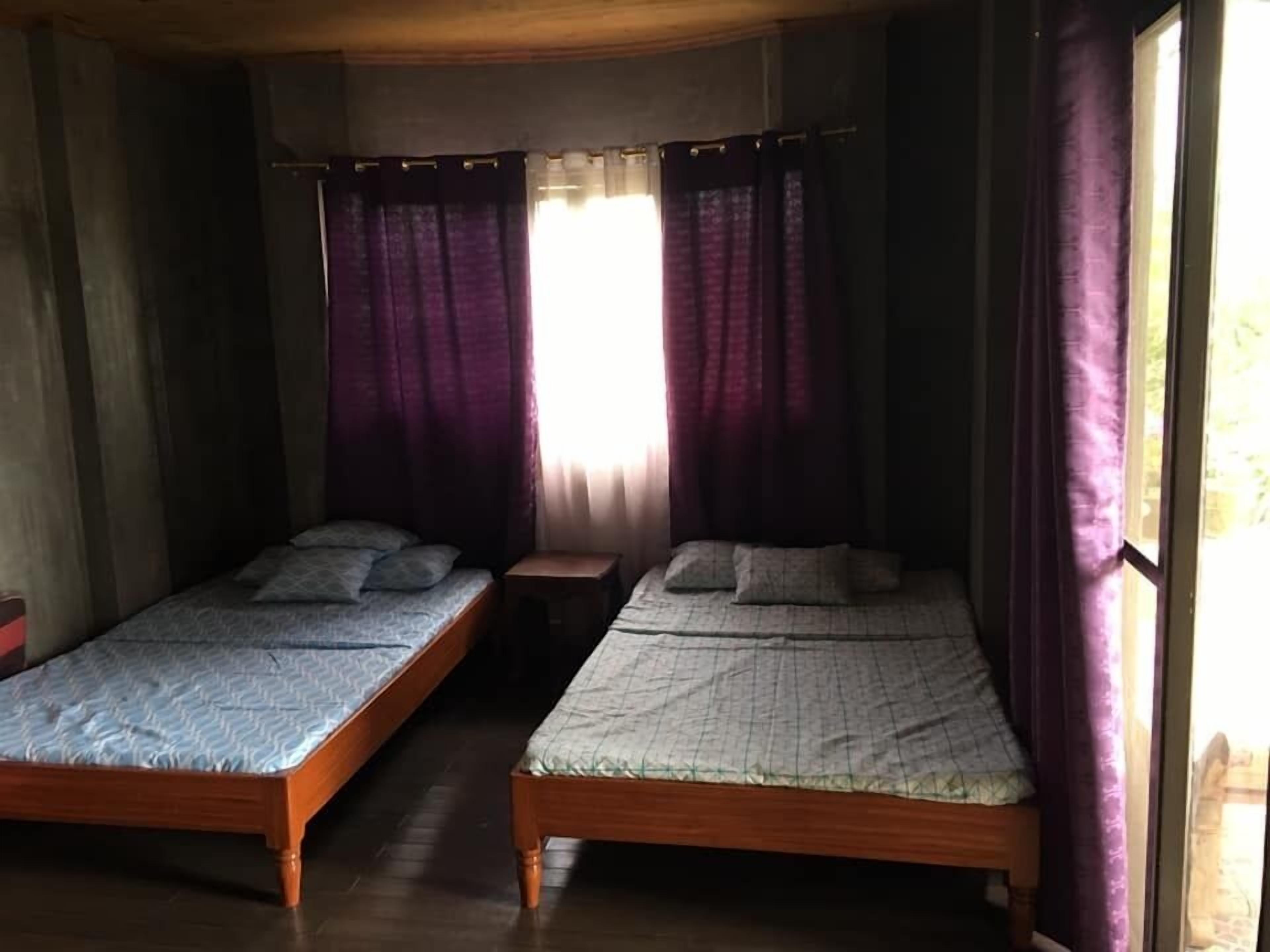 Une chambre avec deux lits en bois, une petite table de chevet en bois et une fenêtre ornée de rideaux violets.