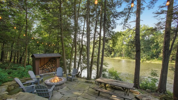 Une terrasse en pierre avec des fauteuils Adirondack, un foyer et un banc en bois donnant sur un lac, le tout entouré de grands arbres.