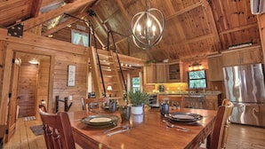 Un intérieur en bois comprenant une table à manger pour quatre personnes, un lustre et une cuisine moderne équipée d’appareils électroménagers en acier inoxydable.