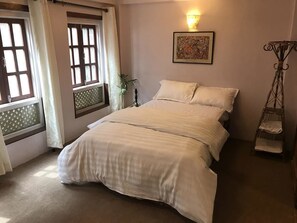 A neatly made bed with white linens in a room with a large window, a wall-mounted light, and a framed picture. - Heritage homes (Lalitpur)