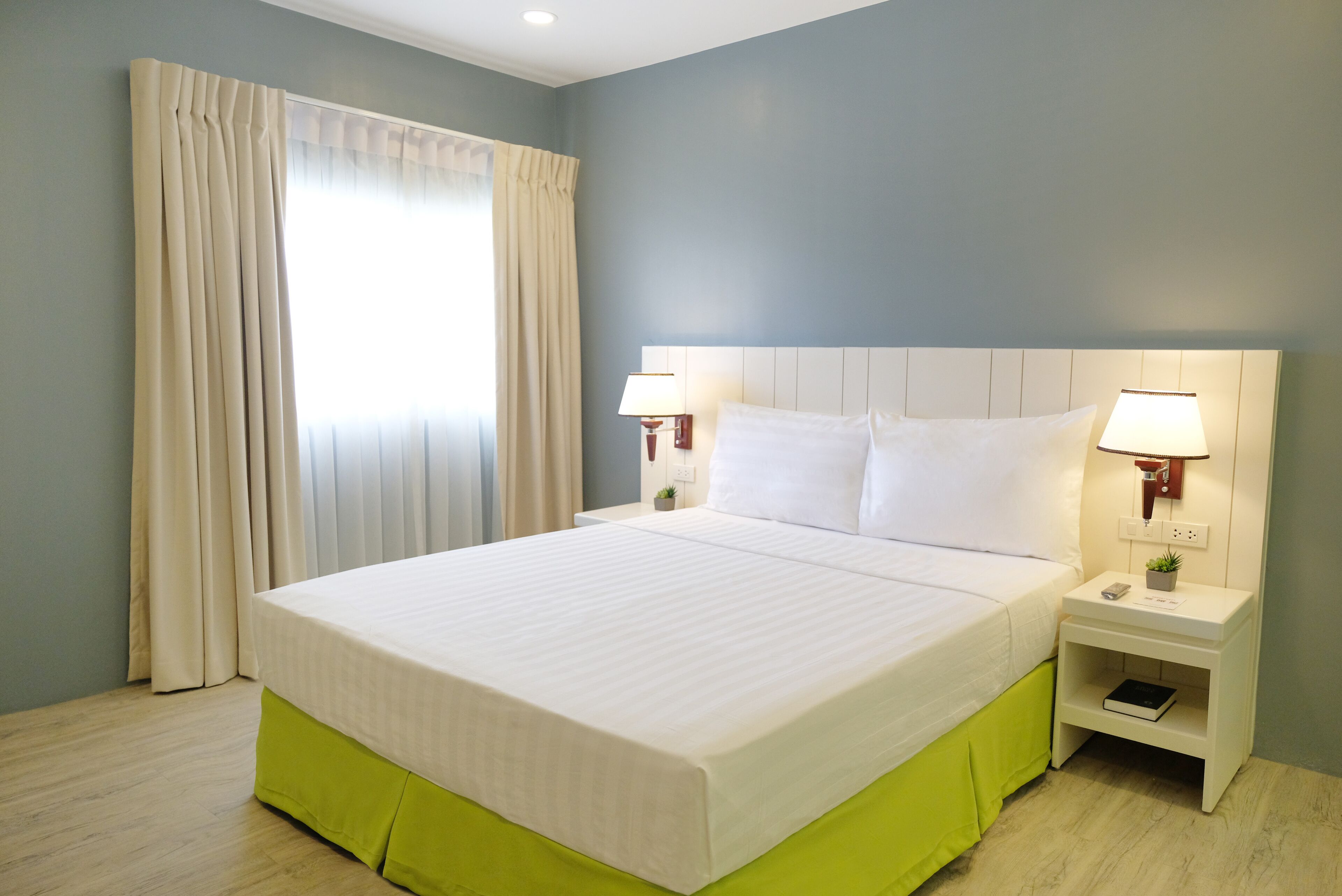 Una cama bien hecha con falda verde, dos mesitas de noche con lámparas y una ventana con cortinas.