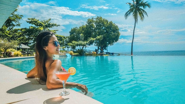 Une personne se détend au bord d’une piscine, un verre à la main, entourée de verdure et sous un ciel dégagé.