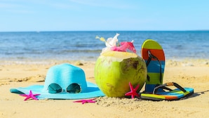 Une scène de plage avec un chapeau bleu, une noix de coco verte munie d’une paille et une paire de tongs colorées sur le sable.