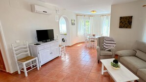 A living room with a sofa, a television, a washing machine, and a dining area with chairs and a table. - 0676 Villa Molins Tauro (Denia)
