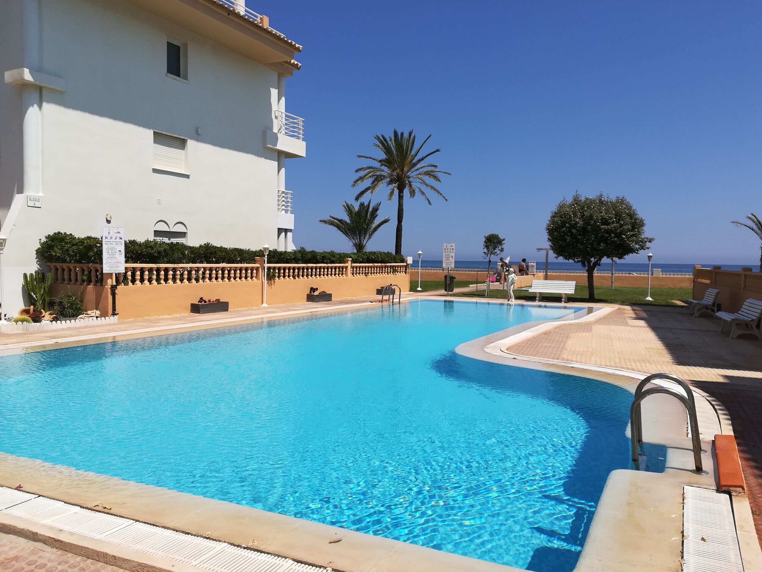 Une piscine à l’eau d’un bleu cristallin, entourée d’un bâtiment blanc doté de balcons, de palmiers et offrant une vue sur la plage.