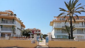 A pathway leading to a beachfront property with multiple-story buildings, a palm tree, and a signboard. - 0770 ap. Talima 1 bedroom (Denia)