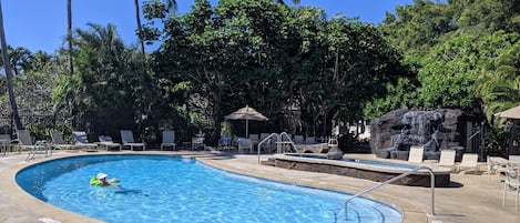 Una piscina con sillas de descanso y una cascada, rodeada de palmeras y follaje.