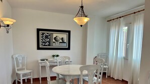Un comedor con una mesa redonda, sillas blancas, un cuadro enmarcado y una ventana con cortinas.