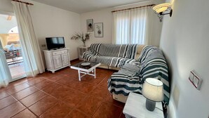 Una sala de estar con un sofá a rayas, un televisor sobre un mueble, una mesa con una lámpara y un ventanal corredizo.