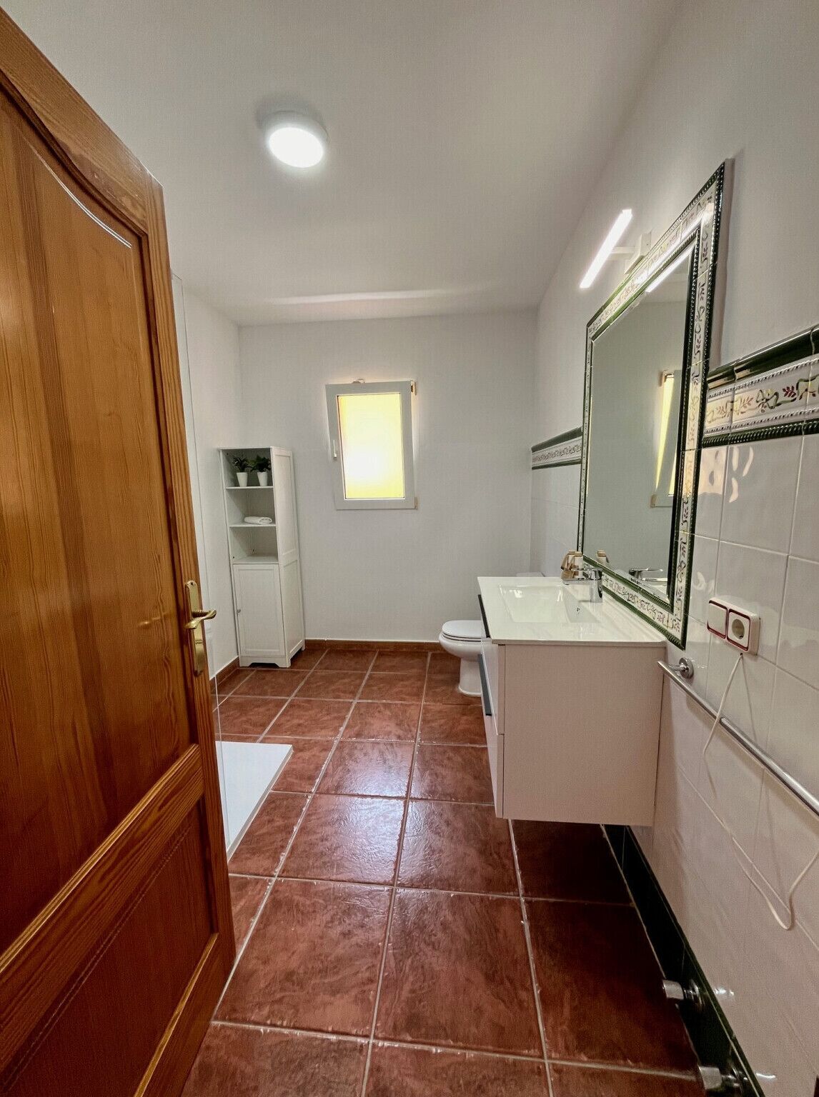 A bathroom with a wooden door, white cabinets, a white sink, a mirror, and a window.