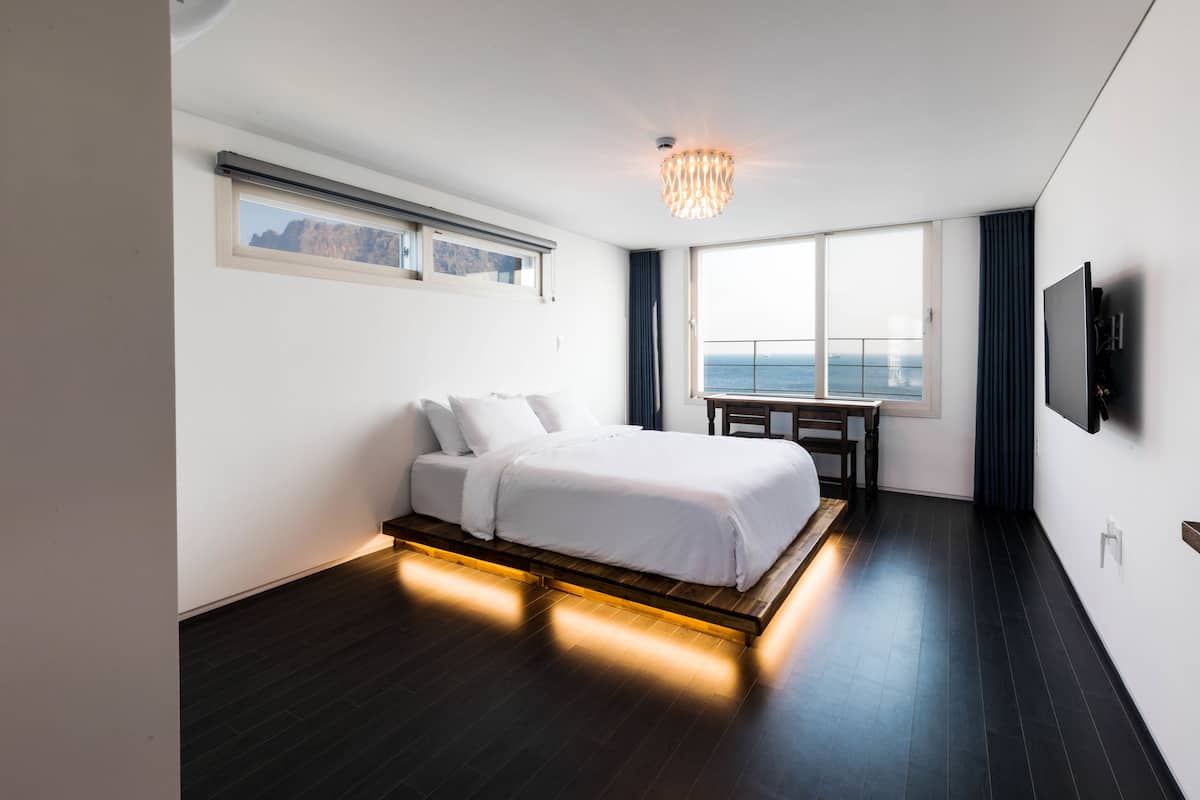 A modern hotel room with a bed, a desk, and a large window offering a view of the sea.