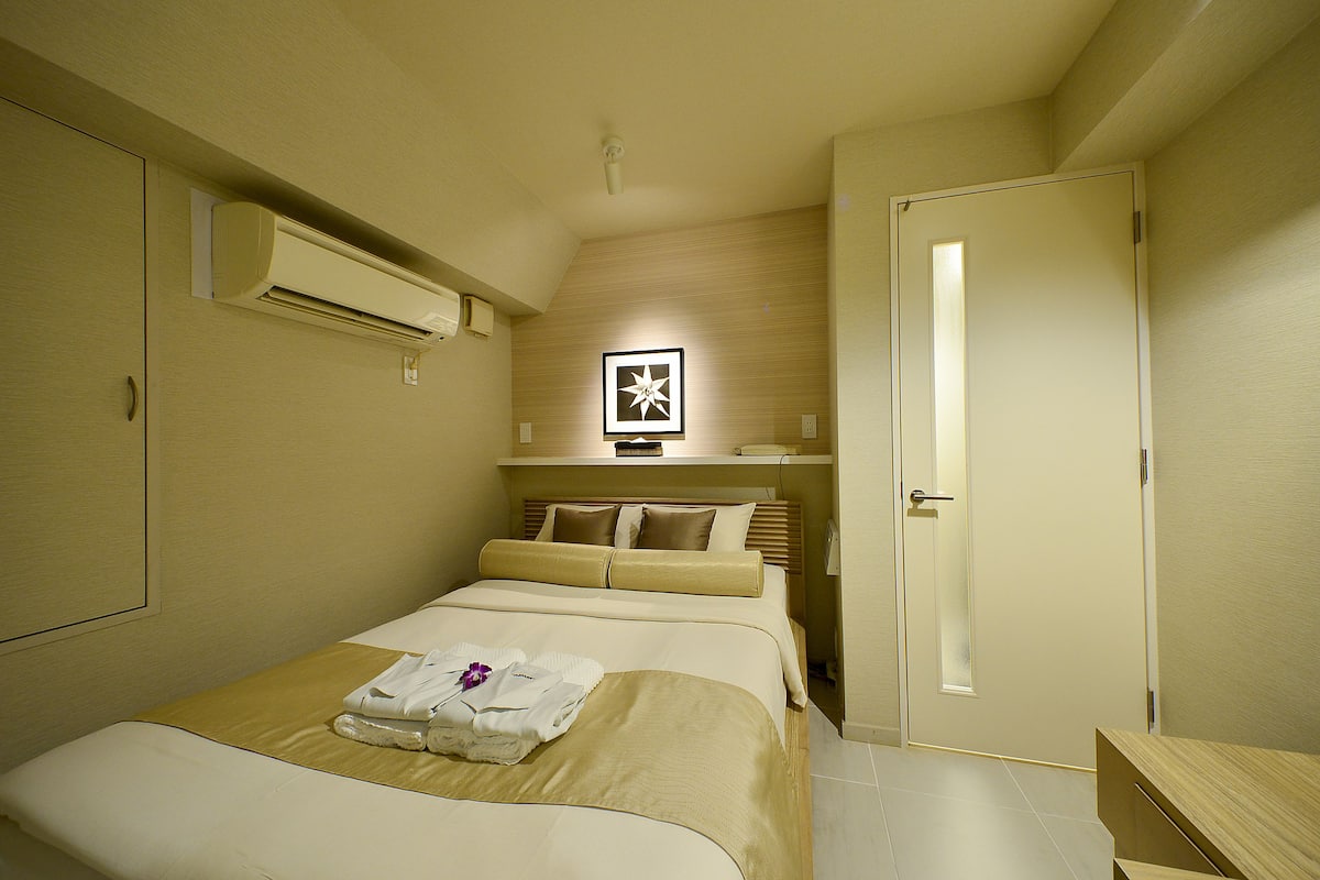 A hotel room with a bed, a bedside table, a wardrobe, and an air conditioning unit.