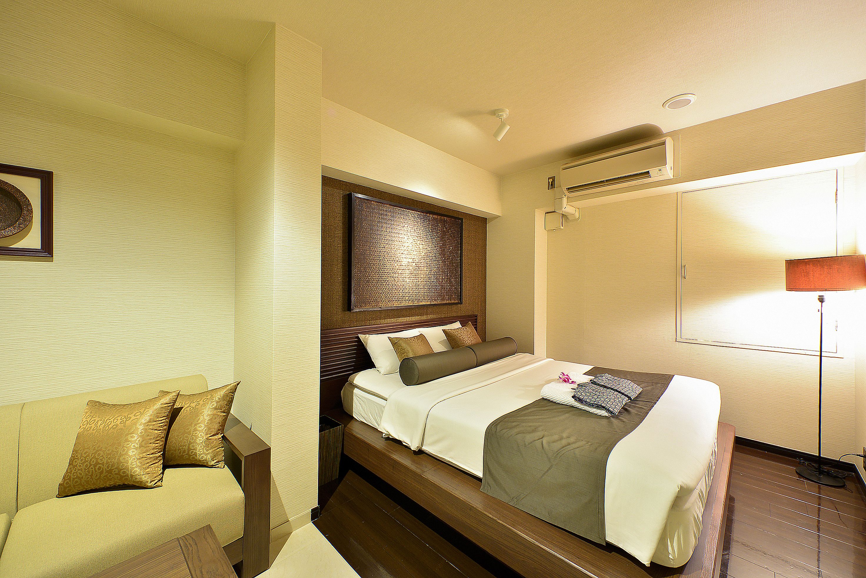A hotel room with a bed, a sofa, a bedside table, and a wall-mounted air conditioner.