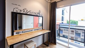 A room with a large mirror, a wooden table with a sign indicating free WiFi and a password, a small stool, and a view of buildings through a window with a curtain. - Mango 10 House (Bangkok)