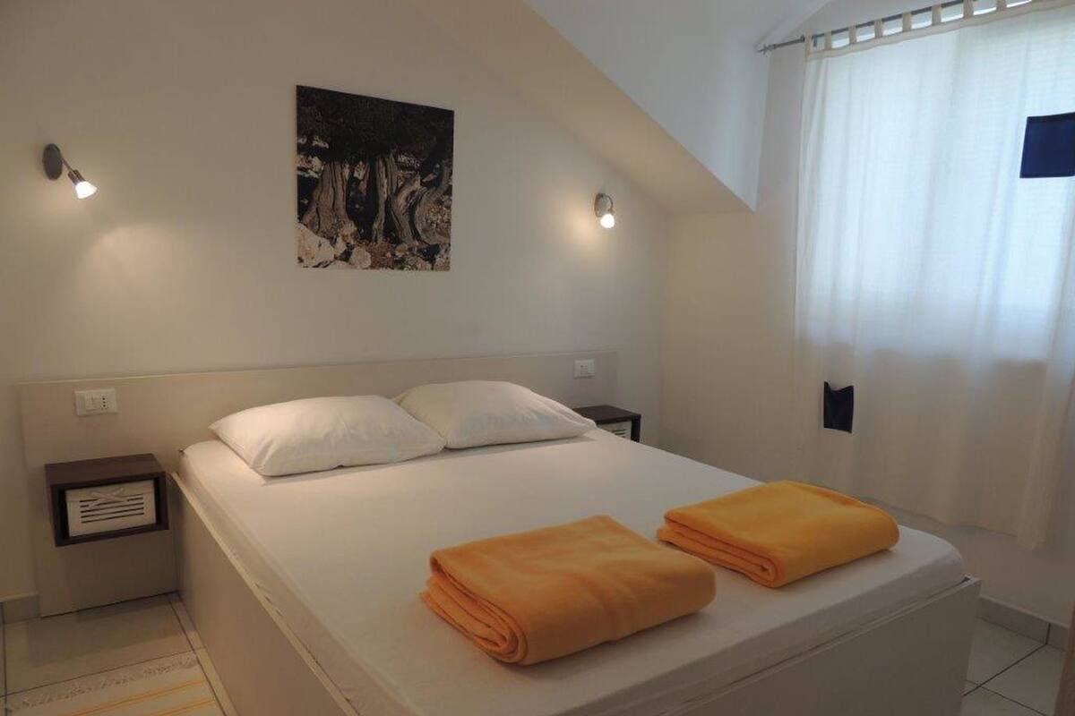 Quarto com cama, duas almofadas, mesa de cabeceira, luminária na parede, quadro e janela com cortinas.