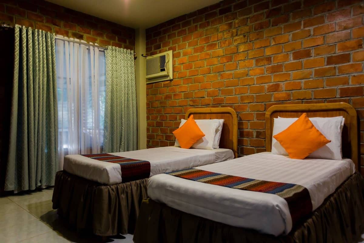 Une chambre d’hôtel avec deux lits, chacun doté d’un matelas blanc et d’un couvre-lit marron, ainsi que d’oreillers orange. Il y a une fenêtre avec des rideaux légers et un mur en briques.