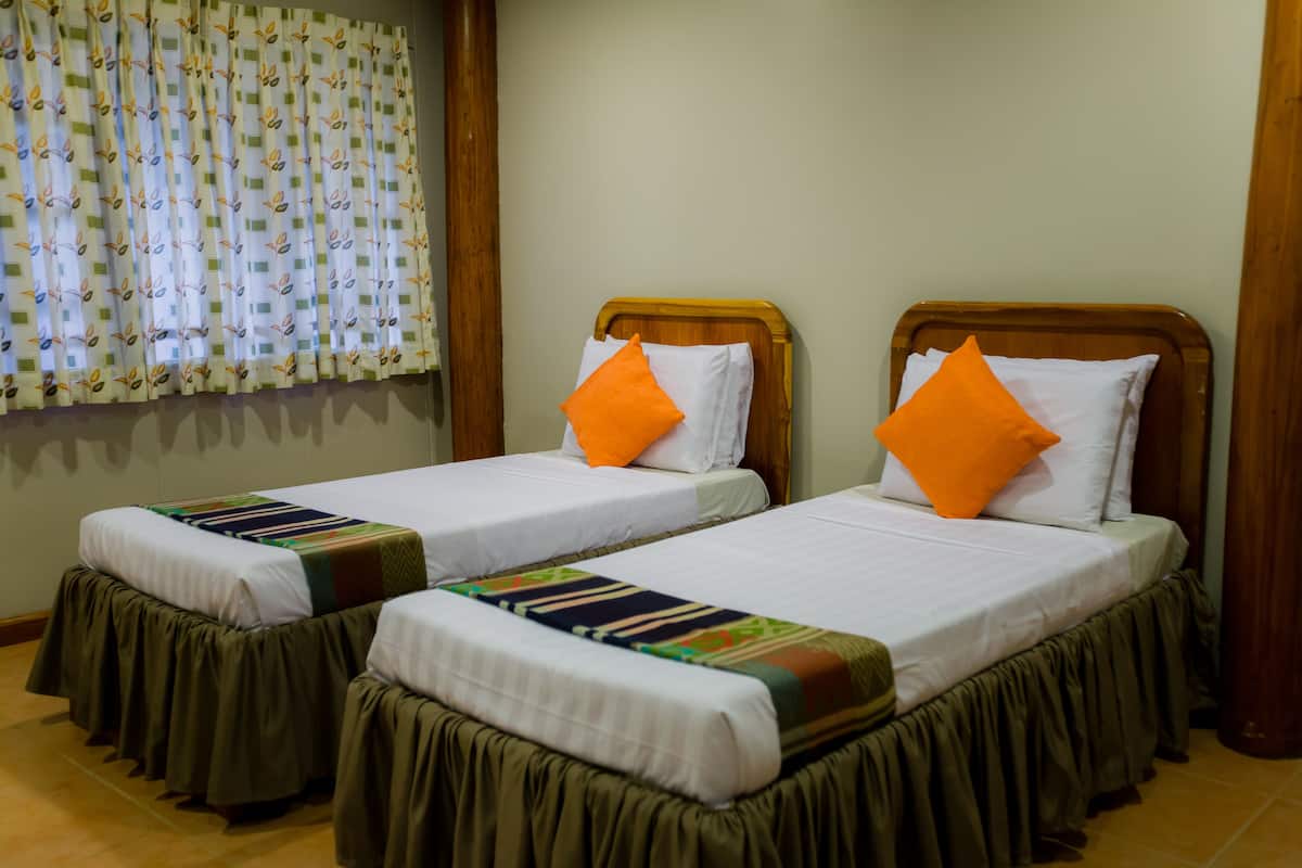 Une chambre d’hôtel avec deux lits simples, chacun doté d’une tête de lit en bois, de draps blancs et d’oreillers orange. La chambre dispose d’une fenêtre avec des rideaux à motifs et d’un plancher en bois.