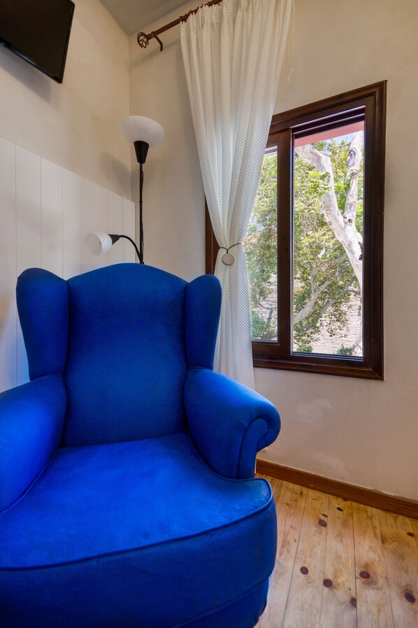 A room with a blue armchair, a floor lamp, a window with a view of trees, and a curtain. - Bastion Medieval Deluxe Apartments (Rhodes)